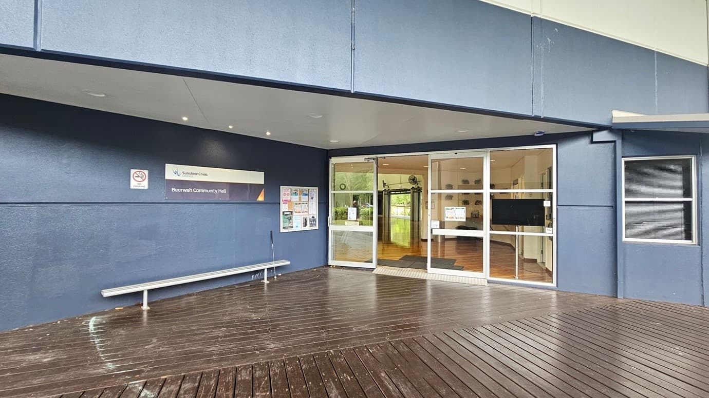 Entrance to a community hall with a blue exterior. On the left, a sign reads "Beerwah Community Hall" above a no-smoking sign. Below, a silver bench is positioned against the wall. A notice board with various posters is nearby. The centre features glass double doors leading inside, revealing a wooden floor and some greenery visible through a window. The right side has a window with closed blinds. The floor is wooden decking, and the ceiling is smooth with recessed lighting.