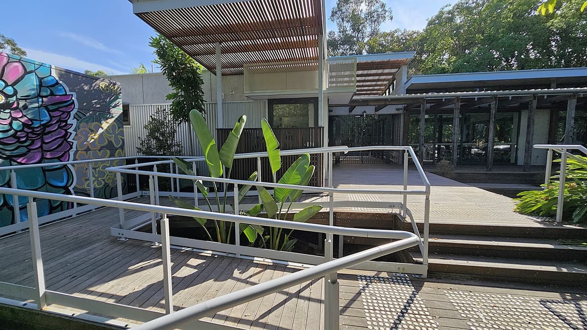 A wooden walkway with metal railings extends in the foreground, surrounded by greenery. On the left, a colourful mural decorates a building wall. In the middle, large green leaves sprout next to a modern building featuring vertical slatted panels and a pergola-like ceiling. On the right, another building with large glass windows overlooks the area. The ground consists of wooden planks, while trees fill the background.
