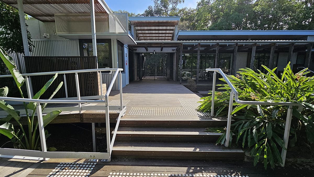 A wooden platform with steps and metal railings forms the central focus, leading to an open corridor. On the left, a building with corrugated metal walls and a slatted overhang adds a modern touch. Large green plants with broad leaves are on the right, flanking the walkway. The floor features textured wooden planks, while the ceiling above the corridor consists of metal and wooden slats. The background is filled with dense, lush trees.