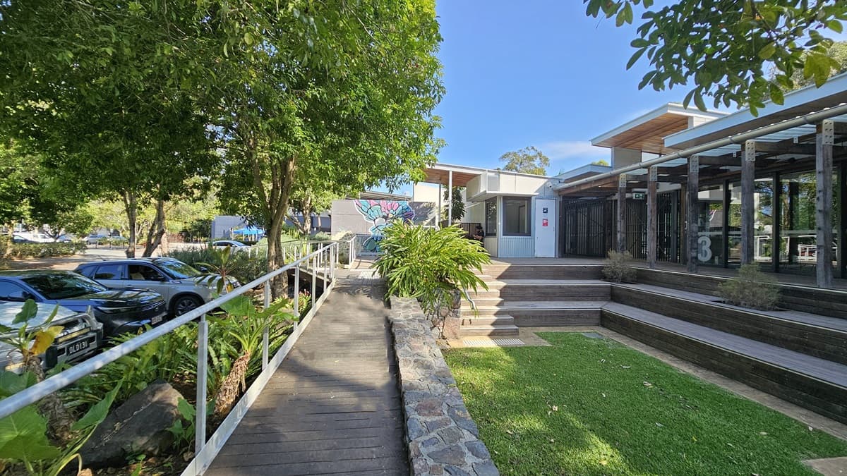 A wooden ramp with metal railings runs along the left, bordered by dense green foliage and parked cars beneath trees. In the middle, steps lead to a modern building with slatted wooden panels and glass sections. A vibrant mural decorates the building's wall. On the right, tiered wooden decking transitions into a grassy area. The ceiling of the building features wooden beams providing partial cover.
