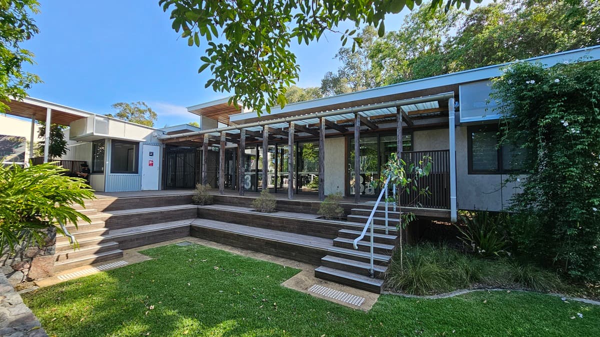 A low, modern building spans the middle of the image, featuring large glass windows and a slatted wooden pergola. On the left, terraced wooden steps lead up to the structure, accompanied by lush green ferns. On the right, a set of stairs with a metal railing descends to a grassy lawn edged with shrubs. Creeping plants cover part of the building. The floor is made of wooden decking, while the ceiling consists of transparent panels. Trees are visible in the background.