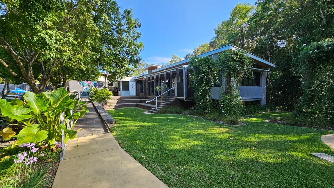 A modern, low-rise building stands in the middle with a slatted facade and vines creeping up the walls. On the left, a well-tended garden features large green leaves and purple flowers beside a concrete path. The right side has a grassy lawn extending to the edge of the building, with trees providing shade. Steps lead up to the entrance, and the building's ceiling is supported by rows of columns. In the background, more trees are visible, creating a secluded, natural setting.