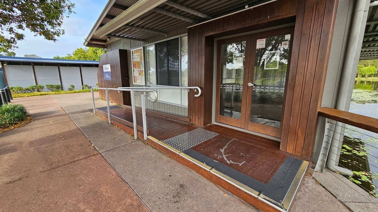 A building with a wooden exterior and glass double doors on the right. A metal handrail runs along a ramp leading to the entrance. The ground is concrete with scattered leaves. On the left, there's a path lined with greenery and a corrugated metal wall in the background. The roof is metal, and a body of water with lily pads is visible on the far right.