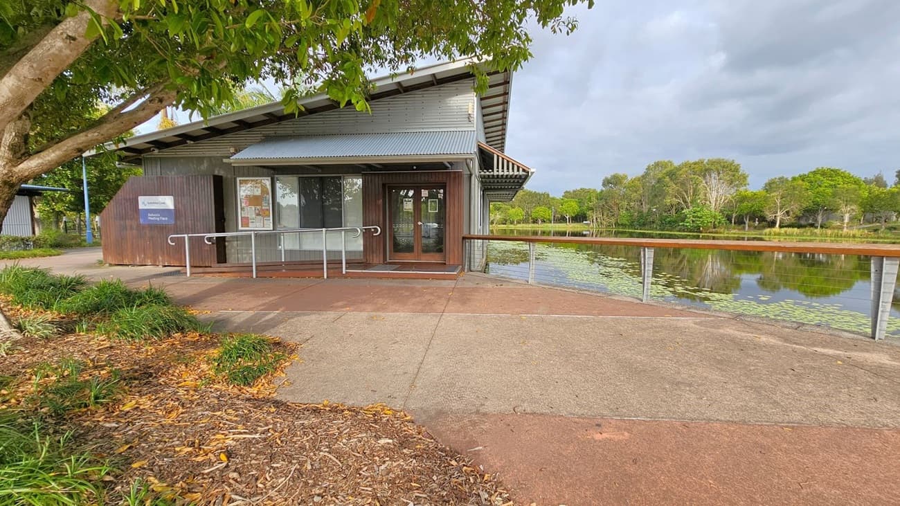 A small building with a metal roof and wooden exterior is on the left, featuring a sign and a glass door. A concrete path leads to the entrance, bordered by mulch and grass. On the right, a wooden railing overlooks a pond with lily pads. Trees line the background, reflecting in the water. A tree with green leaves is in the foreground on the left.