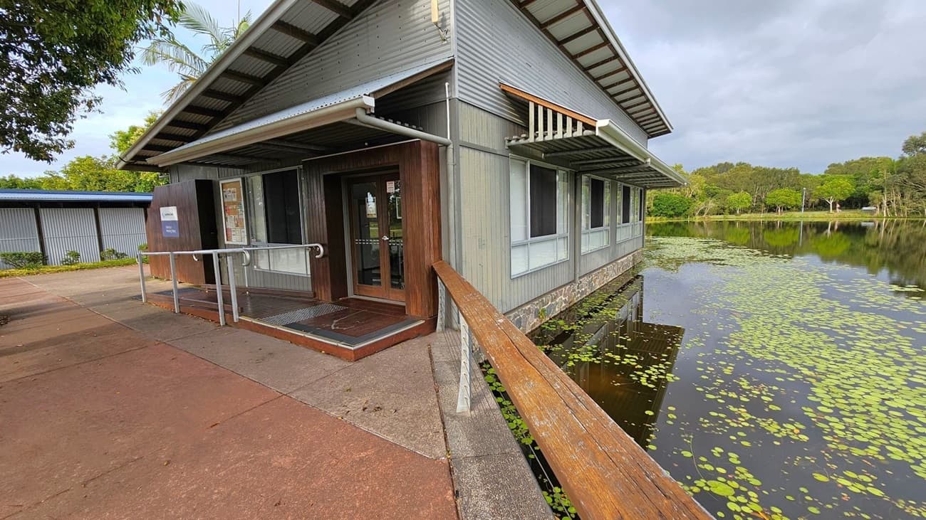 A building with a corrugated metal roof and wooden siding is situated on the edge of a pond covered with lily pads. The entrance features a ramp with a wooden handrail. To the left, a paved walkway leads to another structure with a similar roof. Trees and greenery are visible in the background, reflecting in the water. The building has large windows and a stone foundation.