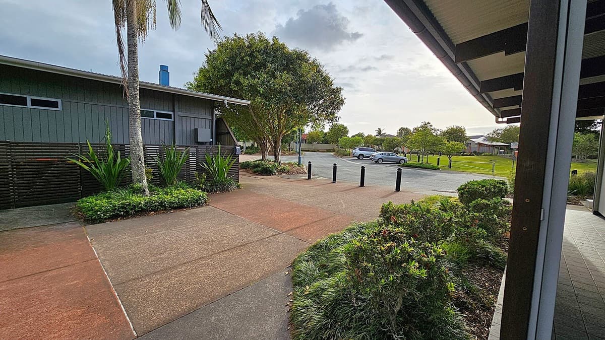A paved walkway with a palm tree and lush green plants on the left, adjacent to a dark wooden building with horizontal windows. In the centre, a large tree stands near a row of black bollards. On the right, a covered walkway with a tiled floor leads to a parking area with a few cars. The background features more trees and a grassy area.