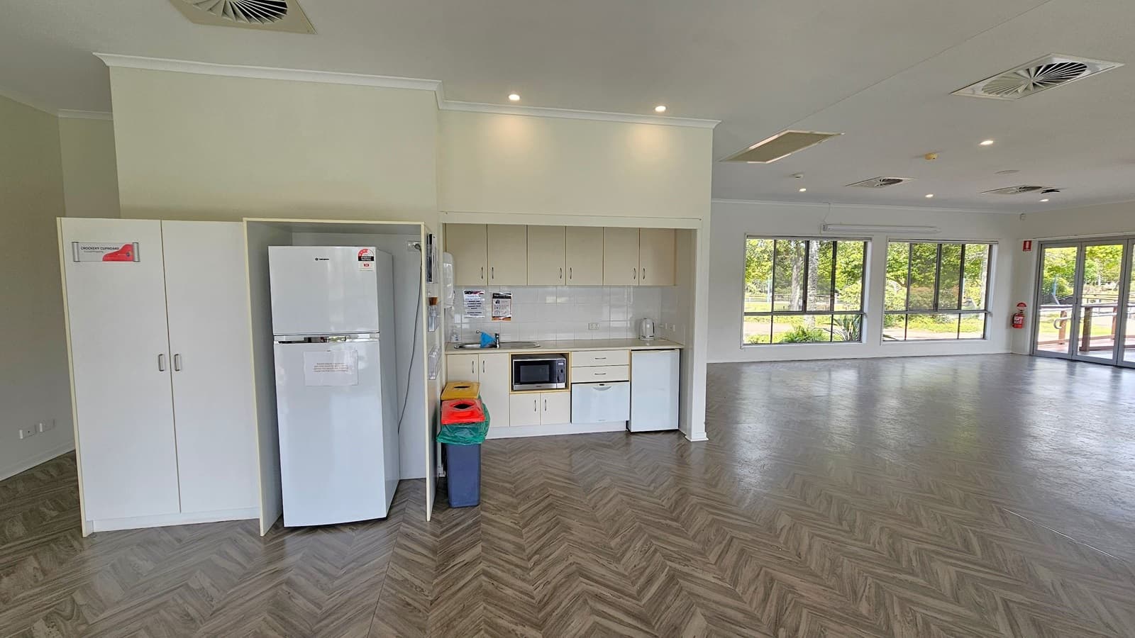 A spacious room with a herringbone-patterned floor. On the left, there is a white cupboard and a refrigerator next to a small kitchenette with cabinets and a sink. The right side features large windows offering views of greenery and a body of water. The ceiling is equipped with recessed lighting and air vents. The far end of the room has glass doors leading outside.