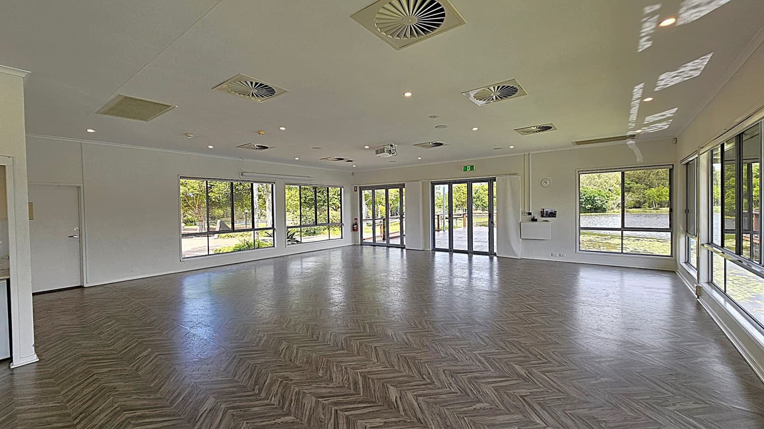 A spacious room with large windows on the right and at the back, offering views of greenery and a body of water. The floor has a herringbone pattern in shades of grey and brown. The ceiling is white with multiple recessed lights and air vents. A set of glass doors is at the back, leading outside. A small wall-mounted shelf and a clock are visible on the back wall.