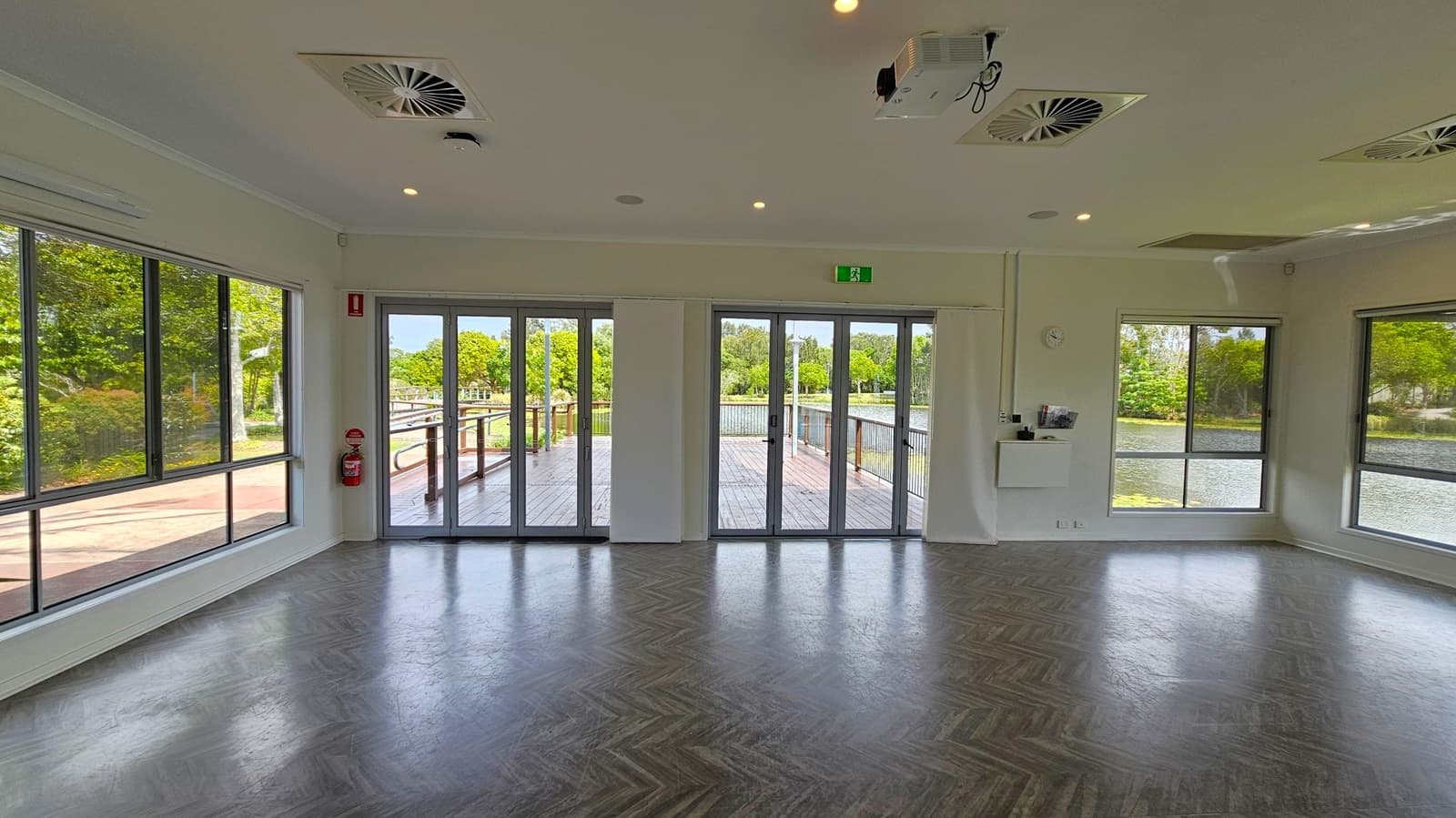A spacious room with large glass doors and windows on the left and right, offering views of a wooden deck and lush greenery outside. The floor has a dark herringbone pattern. The ceiling features recessed lights and air vents, with a projector mounted near the centre. A fire extinguisher is mounted on the left wall, and a small white cabinet with a clock above it is on the right wall.