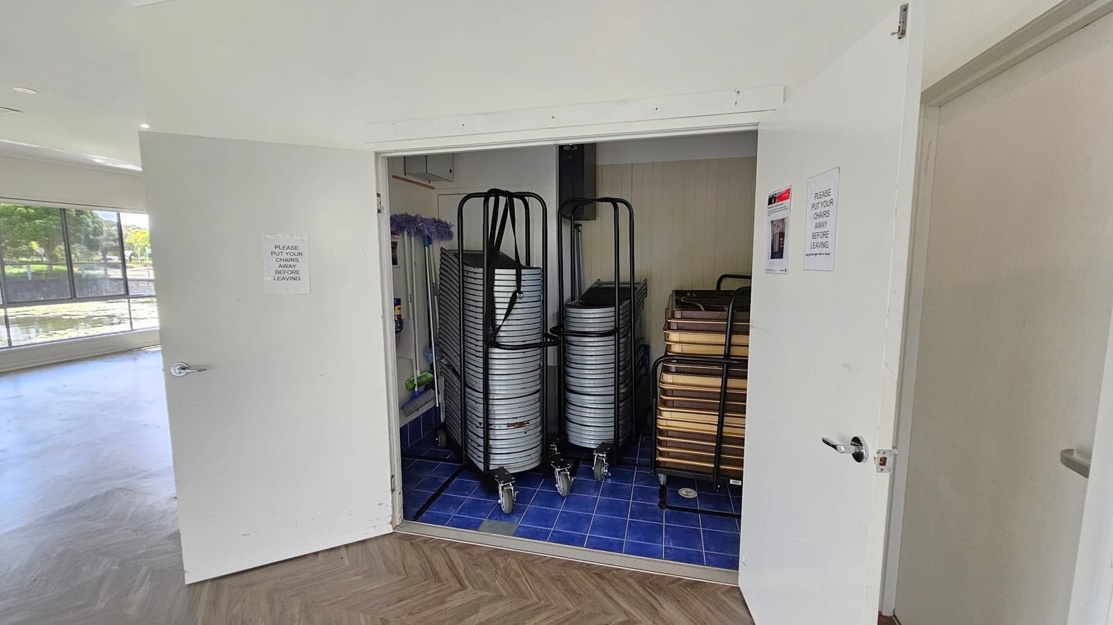 A storage room with open double doors. Inside, there are stacks of metal chairs on trolleys with wheels, and a stack of brown tables on the right. The floor inside the room is covered with blue tiles, while the floor outside is a wood-patterned laminate. The walls are light green. Signs on the doors read, "PLEASE PUT YOUR CHAIRS AWAY BEFORE LEAVING." To the left, large windows reveal a view of greenery and a body of water.