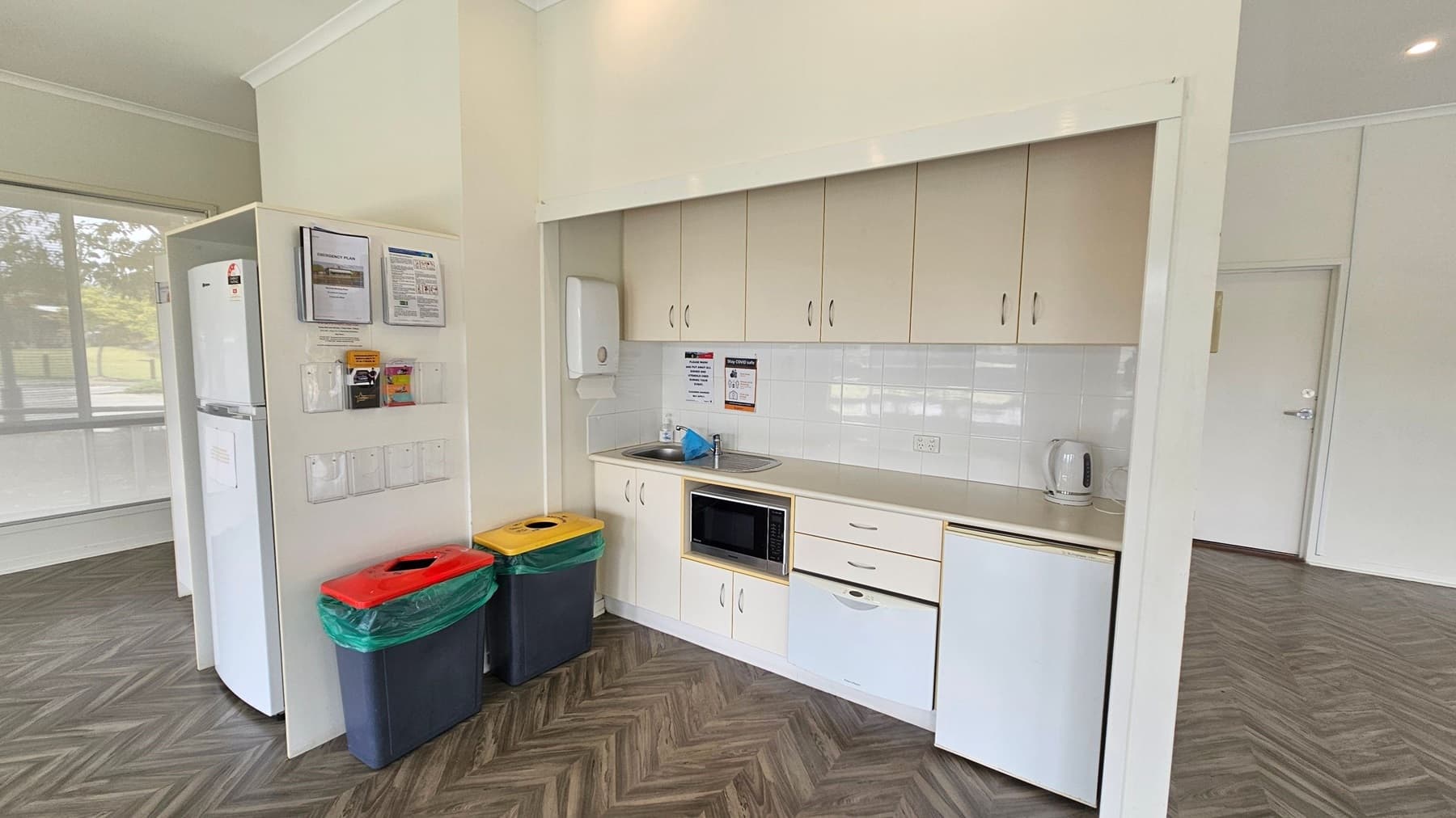 A small kitchen area with light-coloured cabinets and a white tiled backsplash. On the right, there's a countertop with a sink, a microwave, and a kettle. Below are drawers and a mini-fridge. On the left, a white fridge stands next to a wall with notices and brochures. Below the notices are two bins with red and yellow lids. The floor has a herringbone pattern, and the ceiling is plain with recessed lighting. Large windows are visible in the background.
