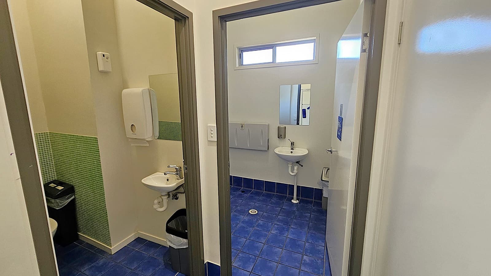 A public restroom with two separate stalls. The left stall has a small sink, a soap dispenser, and a waste bin. The walls have green tiles halfway up. The right stall features a similar sink and soap dispenser, a hand dryer, and a mirror. The floor is covered with blue tiles, and there is a small window near the ceiling.