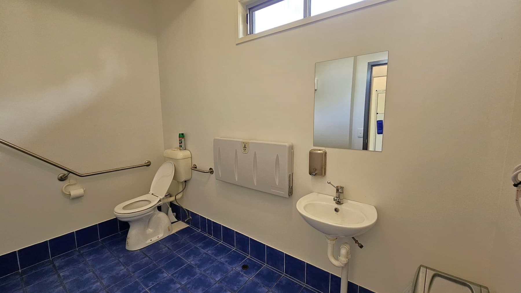 A bathroom with blue-tiled flooring and white walls. On the left, there is a toilet with a grab rail and a toilet roll holder. Above the toilet is a small window. In the centre, a baby changing station is mounted on the wall. On the right, a small sink with a soap dispenser is below a rectangular mirror. A metal bin is partially visible in the lower right corner.