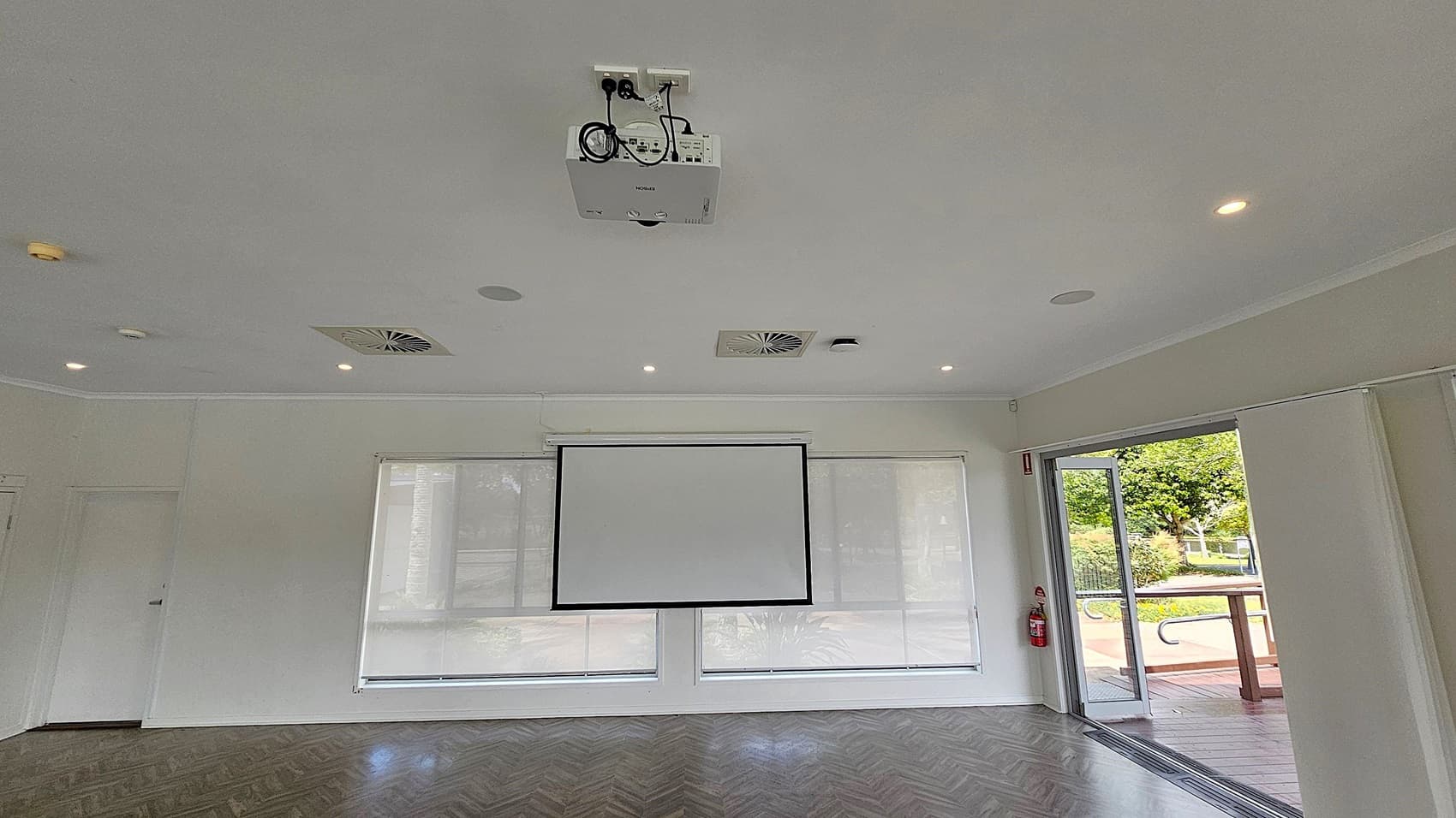 A room with a white ceiling featuring recessed lights and air vents. A projector is mounted centrally. The back wall has a large projection screen flanked by two windows with blinds. On the right, a glass door is open, leading to an outdoor area with greenery. A fire extinguisher is mounted near the door. The floor has a dark herringbone pattern.