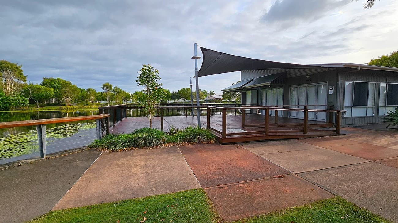 A wooden deck with a ramp and metal handrails extends from a grey building on the right. The building has large windows. Above the deck, a dark triangular shade sail is suspended. In the background, there is a pond surrounded by lush green trees. The foreground features a multicoloured concrete path with patches of grass.