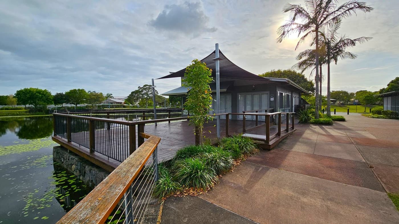 A modern building with a shaded deck is situated by a pond on the left, with lily pads floating on the water. The deck is surrounded by a black metal railing and has a small tree and greenery at its base. On the right, a concrete path leads past the building, flanked by tall palm trees and more greenery. In the background, there are more trees and a grassy area. The sky is overcast.