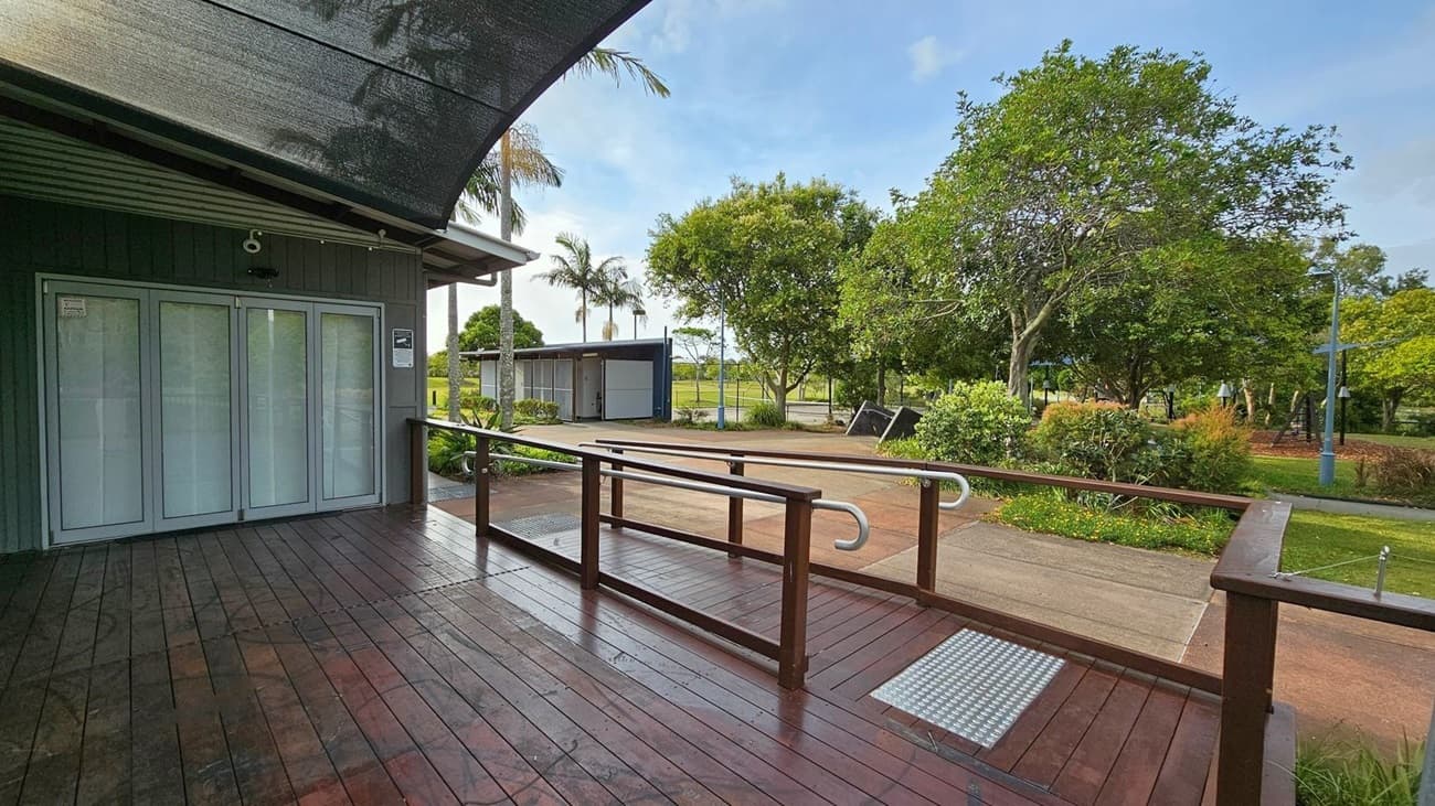 A wooden deck with a ramp and handrails is in the foreground, leading to a building with light blue doors on the left. The deck is covered by a dark canopy. In the background, there are trees, a small building, and a pathway. The area is surrounded by greenery, including bushes and a few palm trees. A lamppost is visible on the right side near the trees.