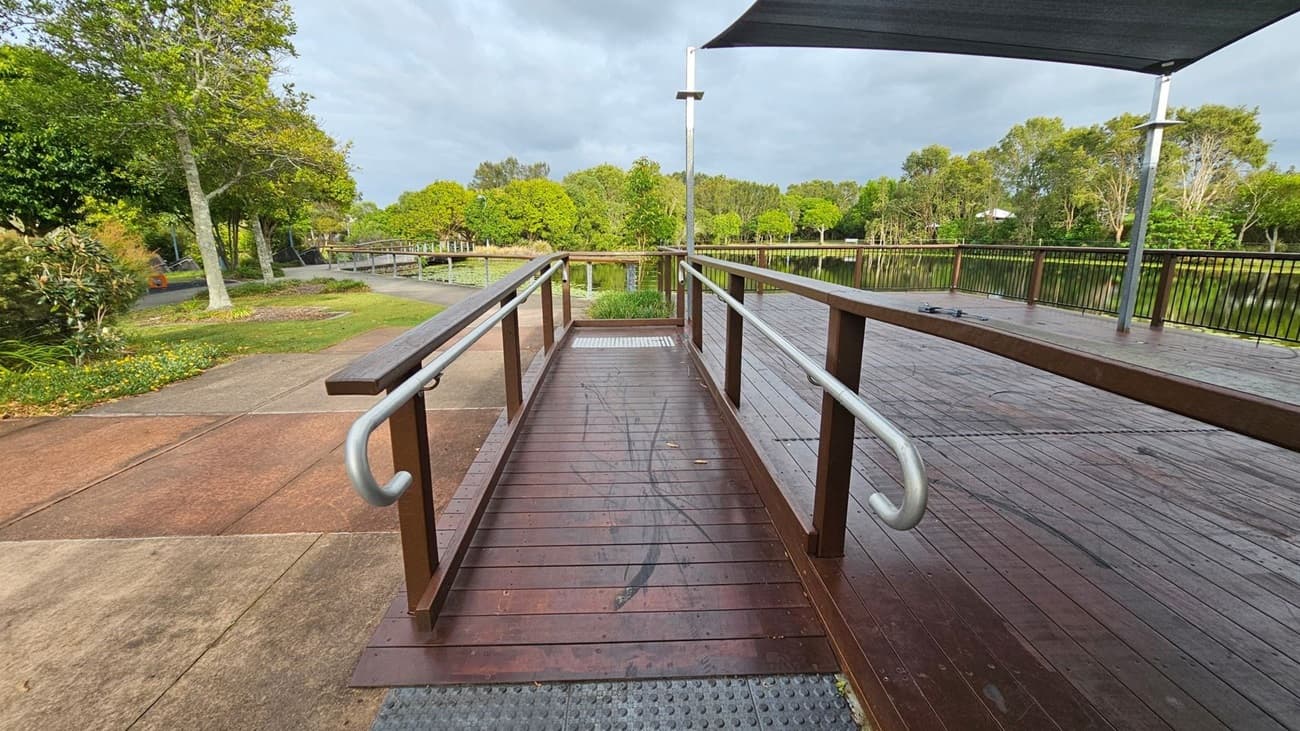 A wooden ramp with metal railings leads to a deck on the right. The deck is covered by a canopy and bordered by a metal fence. On the left, there's a paved path surrounded by green grass and trees. In the background, a pond is visible, lined with more trees and vegetation. The ground is a mix of concrete and wooden planks.