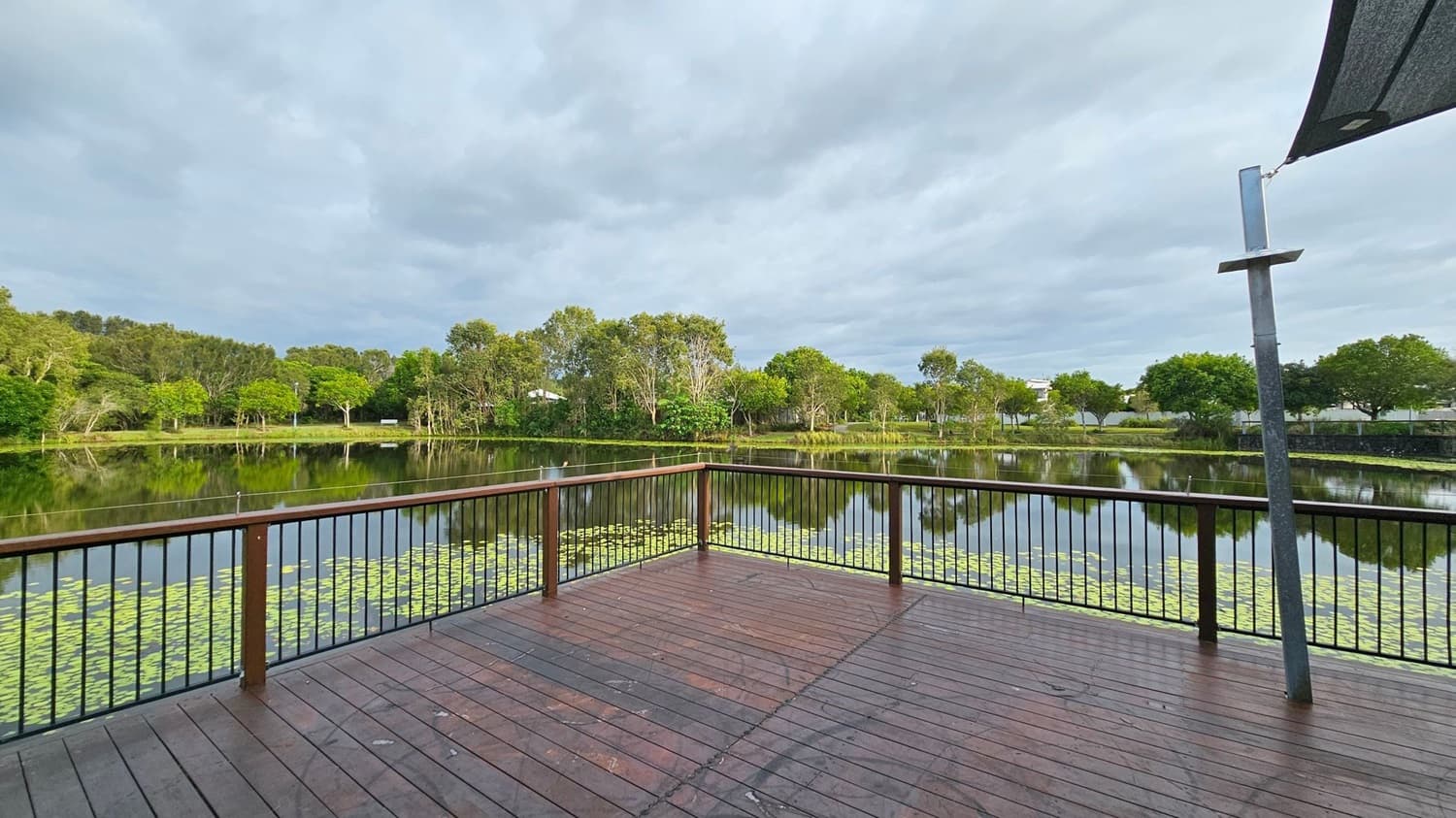 A wooden deck with a railing overlooks a calm lake covered with lily pads. The lake reflects the surrounding lush green trees. On the right, a metal pole supports a partial canopy. The background features a line of trees and some distant structures. The deck's surface is slightly weathered, and the scene is tranquil.