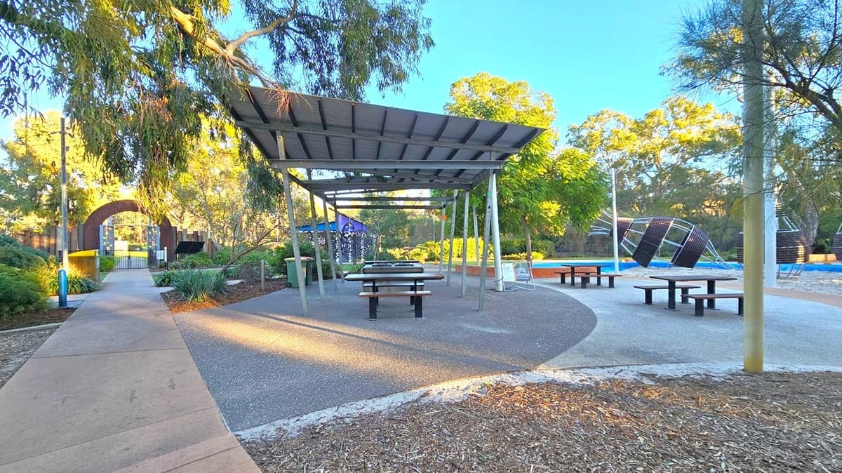 A park scene with a covered picnic area in the centre, featuring a metal roof and picnic tables beneath. To the right, there are modern playground structures on a blue surface. On the left, a pathway leads to a gated entrance with colourful panels. The area is surrounded by trees and greenery, with wood chips on the ground in the foreground.