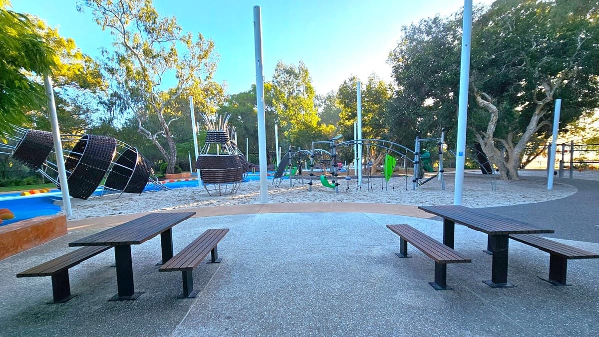 A playground with modern climbing structures made of metal and dark materials is in the background, surrounded by sand. Tall white poles are scattered throughout the area. In the foreground, there are two wooden picnic tables with attached benches on a speckled concrete surface. Trees with dense foliage are visible in the background, providing shade and a natural setting.