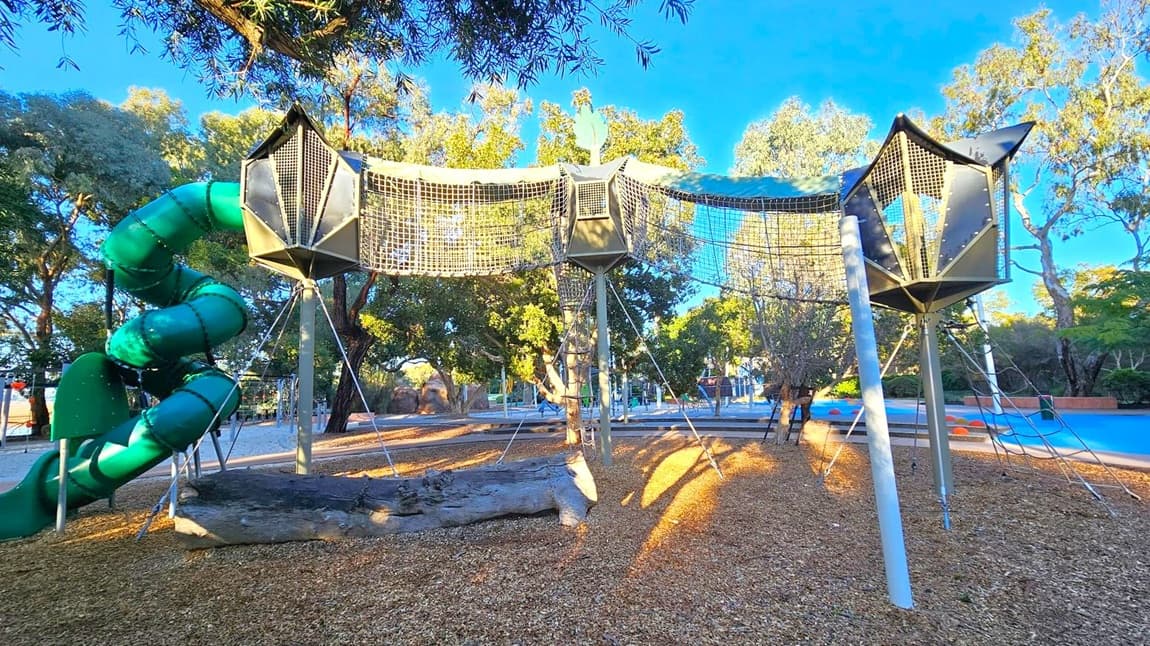 A playground with a large, green spiral slide on the left. In the centre, there are elevated netted walkways supported by metal poles, connecting three geometric structures. The ground is covered with wood chips, and there are trees in the background providing shade. To the right, a blue play area is visible with orange stepping stones. The scene is well-lit, suggesting it's daytime.