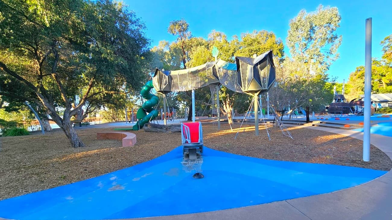 A playground with a variety of equipment. In the centre, there is a netted climbing structure supported by poles. To the left, a large green spiral slide is visible, partially obscured by trees. The ground is covered with wood chips and blue rubber flooring. On the right, there are more play structures, including a spherical climbing frame. Trees surround the area, providing shade, and a curved bench is visible on the left. The sky is clear and blue.