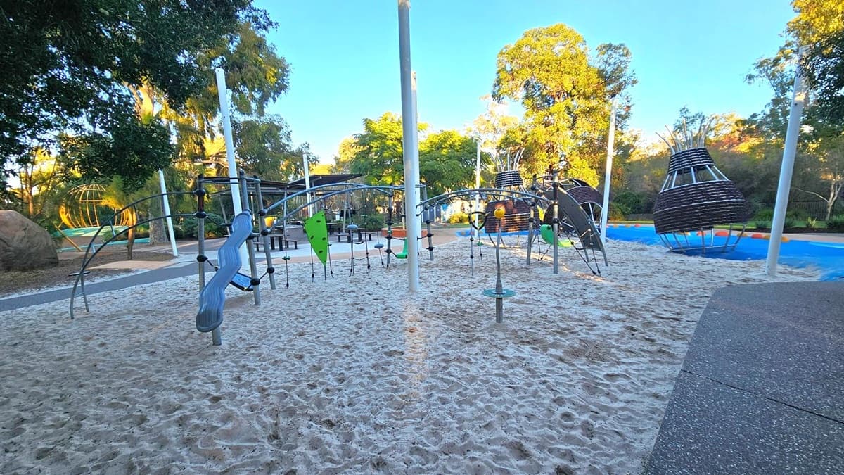 A playground with sandy ground features various climbing structures and play equipment. In the centre, there are metal frames with ropes and climbing panels. To the right, there are large, basket-like structures on a blue surface. Trees and greenery surround the area, providing shade. A pathway with a textured surface runs along the right side.