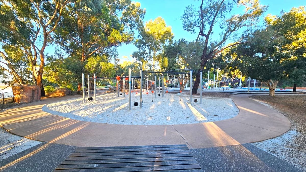 Playground with various climbing structures in the centre, surrounded by sand. A curved pathway encircles the play area. Tall trees with dense foliage are in the background, providing shade. The ground is a mix of sand and paved surfaces. A bench is visible in the foreground.