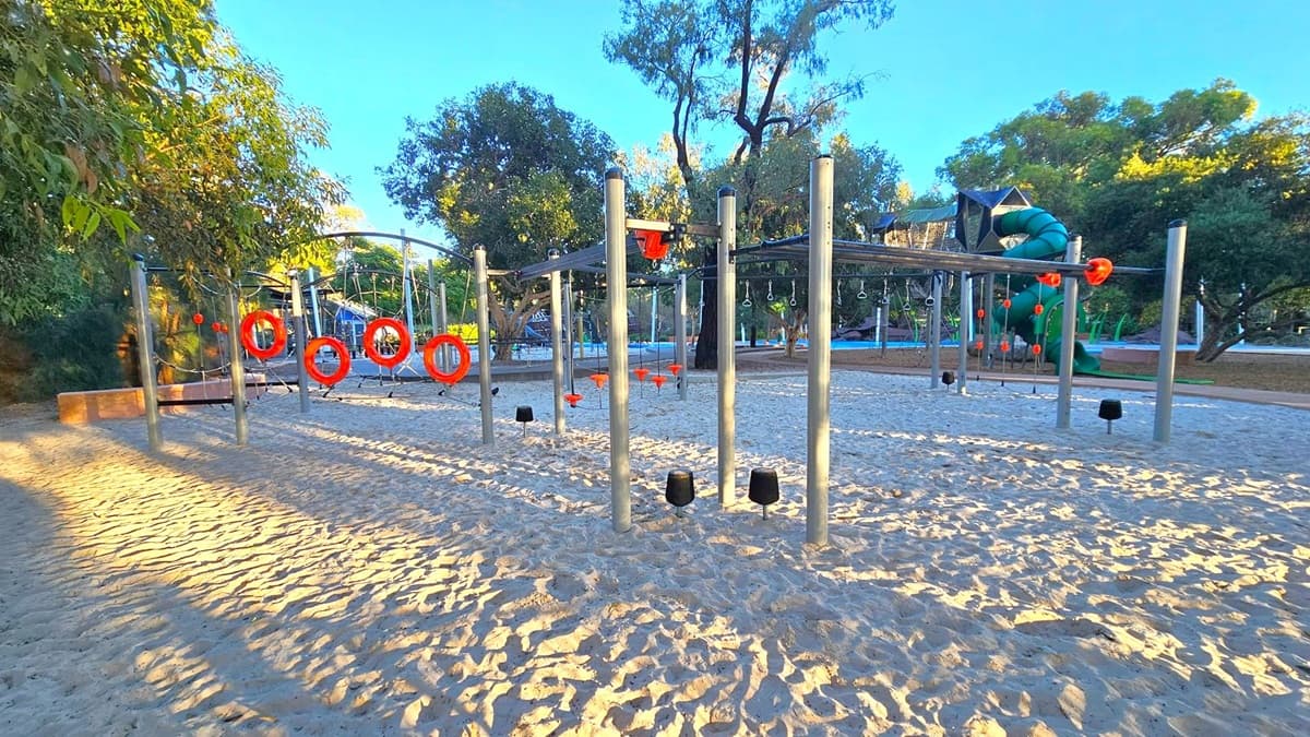 A playground with sandy ground features a climbing structure with metal poles and orange circular elements. On the left, there are trees providing shade. In the background, a large green spiral slide is visible, along with more playground equipment. The area is surrounded by more trees, creating a natural setting. The lighting suggests it's either early morning or late afternoon, casting long shadows across the sand.