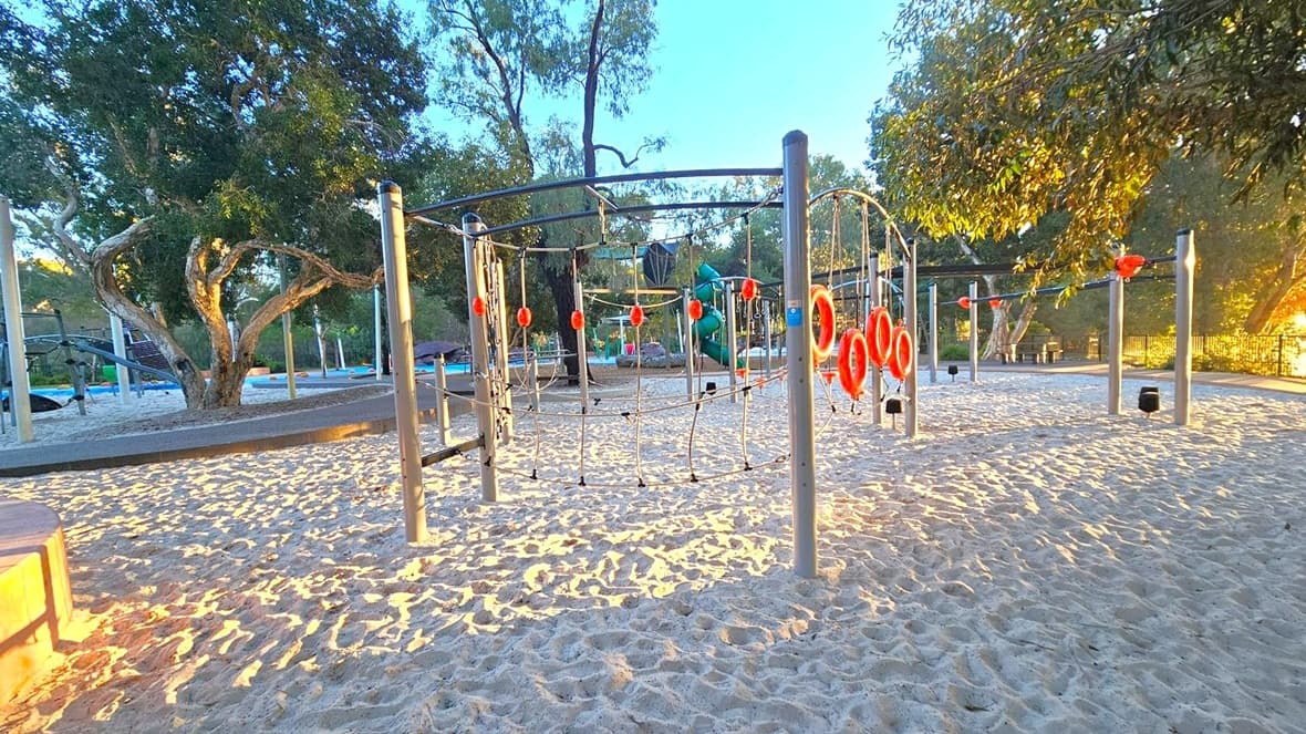A playground with sandy ground featuring a climbing structure with metal poles and ropes, adorned with orange rings. On the left, a large tree provides shade, while more playground equipment is visible in the background. To the right, another tree with dense foliage is present. The area is surrounded by greenery, and the lighting suggests a late afternoon setting.