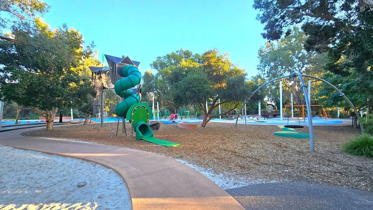 A playground with a green spiral slide structure on the left, surrounded by wood chips. A curved path runs through the area, bordered by sand on the left. In the middle, there are various play equipment, including a swing set with a circular swing. Trees with dense foliage are scattered throughout, providing shade. The ground is a mix of sand, wood chips, and rubberised surfaces.