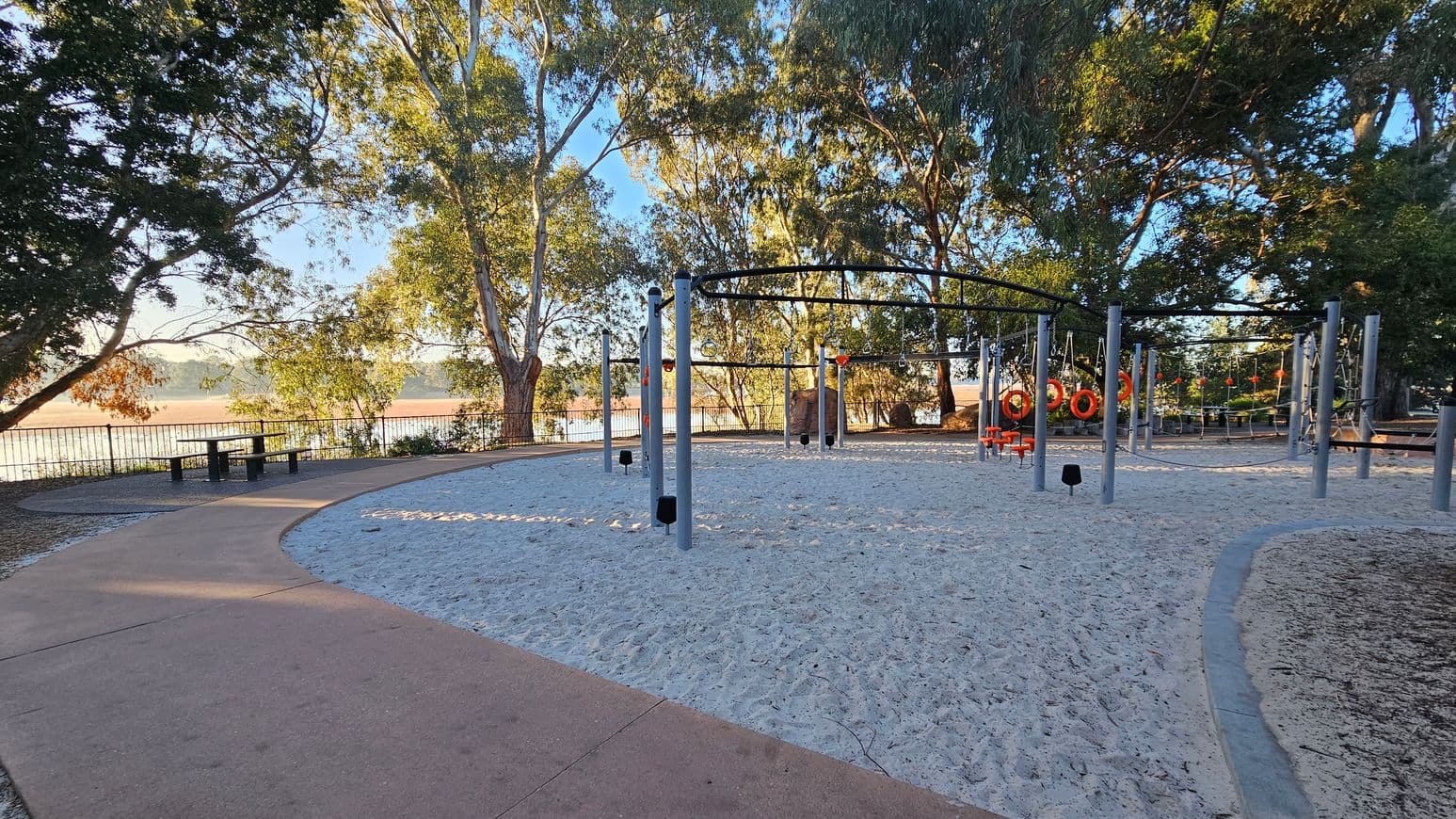 A playground with sandy ground and various climbing structures and equipment, including rings and poles, is in the centre. On the left, a curved path leads to a picnic table under tall trees. The background features a metal fence with a view of a body of water and more trees. The area is shaded by the surrounding trees, creating a mix of light and shadow on the ground.