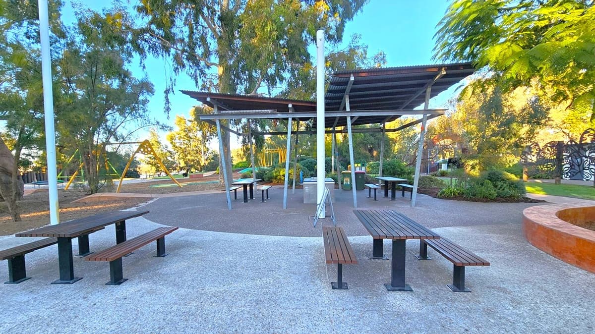A park setting with wooden picnic tables and benches on a paved surface in the foreground. In the middle, there's a covered area with a corrugated metal roof supported by metal poles, housing more tables and a barbecue station. In the background, a playground with yellow climbing structures is visible, surrounded by trees and greenery. A decorative metal fence is on the right, and the ground is a mix of concrete and mulch.