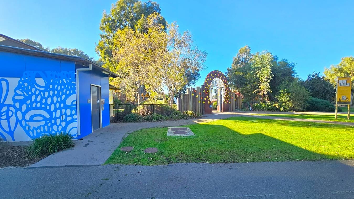 A blue building with a white abstract pattern is on the left, featuring a door and some plants at its base. In the centre, a path leads to an arched entrance decorated with yellow shapes, surrounded by trees and greenery. On the right, a sign reads "Report a Problem" next to a grassy area. The background is filled with tall trees under a clear blue sky.