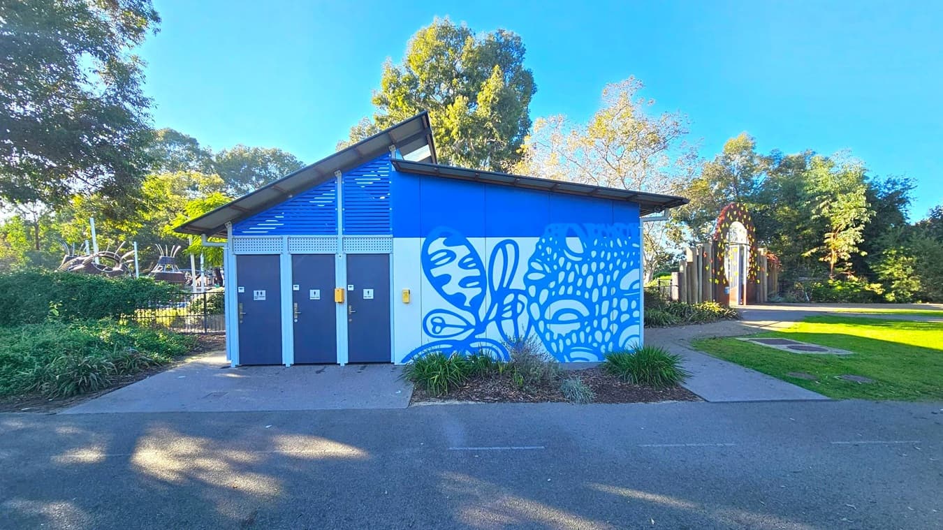 A small building with a slanted roof, featuring three dark grey doors on the left, each with signage. The right side has a blue and white mural with abstract patterns. Surrounding the building are green plants and trees. A pathway leads to a colourful arched gate on the right. The ground is a mix of concrete and grass, with shadows from the trees.