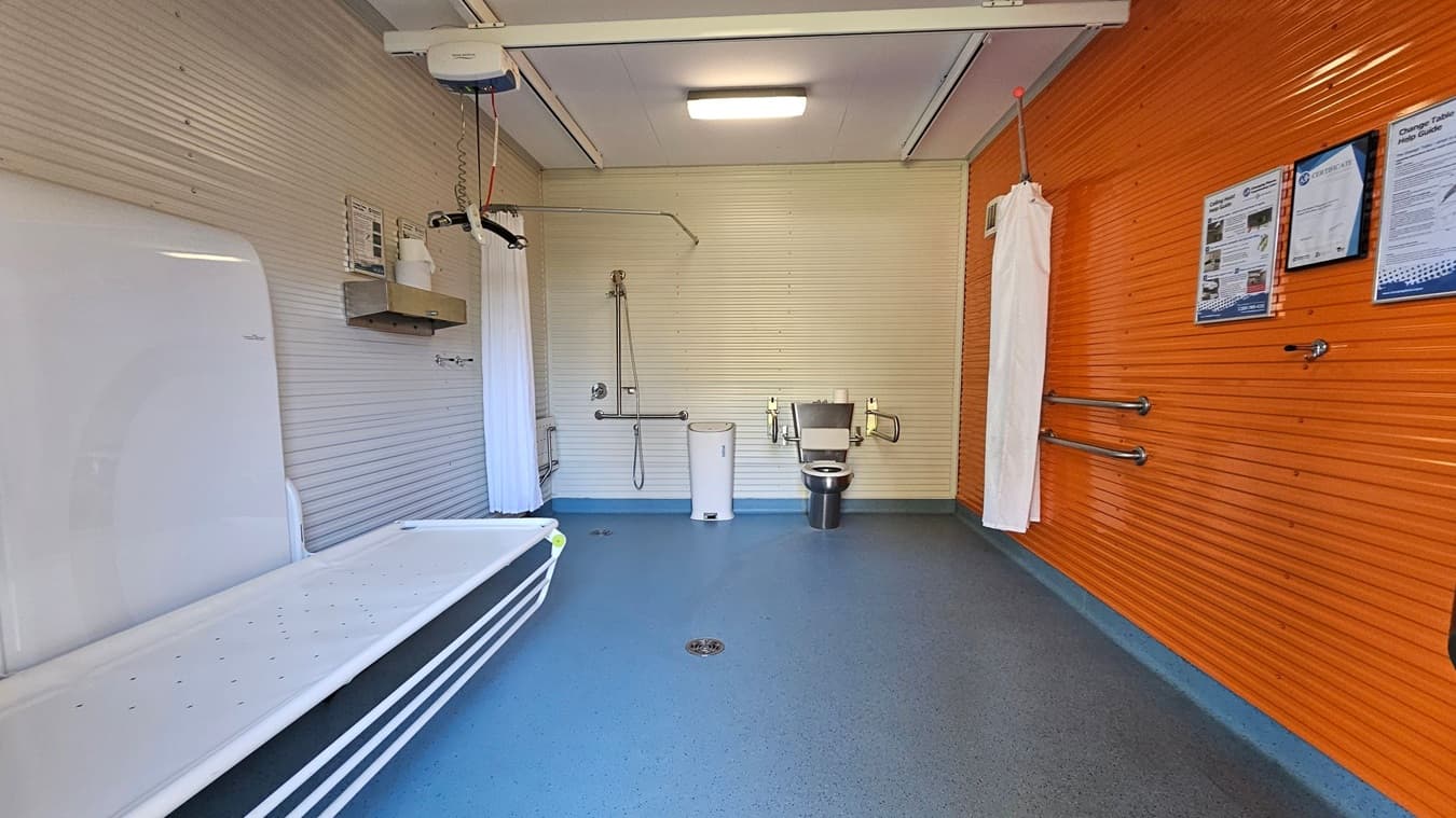 A spacious, accessible bathroom with a blue speckled floor and a white ceiling. On the left, there's a foldable changing table. The back wall features a shower area with a handheld showerhead and grab bars, alongside a stainless steel toilet with support rails. A white bin is positioned between them. The right wall is bright orange with informational posters and additional grab bars. A ceiling track hoist is also visible.