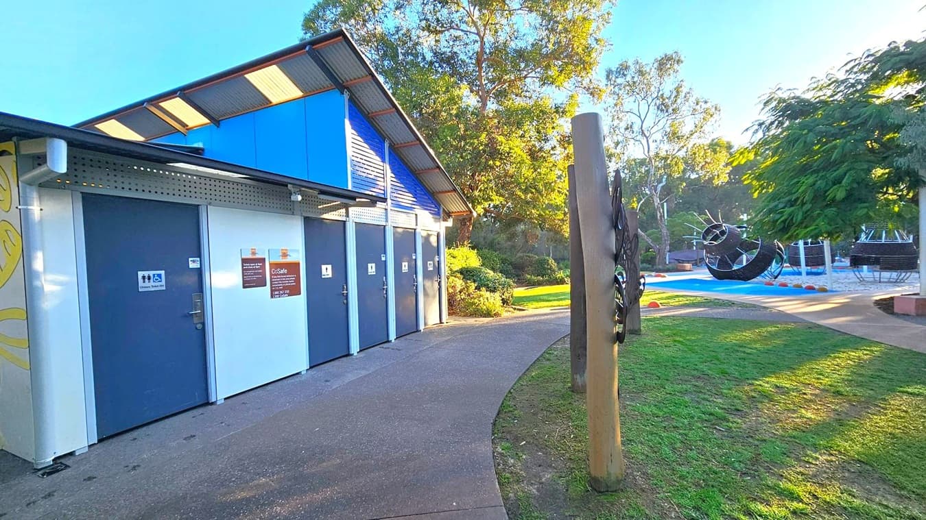 A public restroom facility with blue doors and a corrugated metal roof is on the left. Signs indicate accessibility features. A paved path curves around the building, leading to a grassy area with trees in the background. On the right, a playground with large, spherical climbing structures is visible. A wooden post with decorative elements stands near the path. The area is well-lit, suggesting a sunny day.