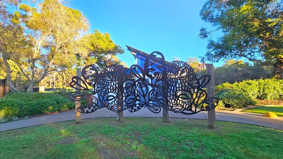 A decorative metal fence with intricate, abstract patterns stands in the foreground, supported by wooden posts. Behind it, a modern building with a slanted roof is partially visible. The background features lush greenery, including trees and shrubs, under a clear blue sky. The ground is covered with grass and a paved path runs alongside the fence.