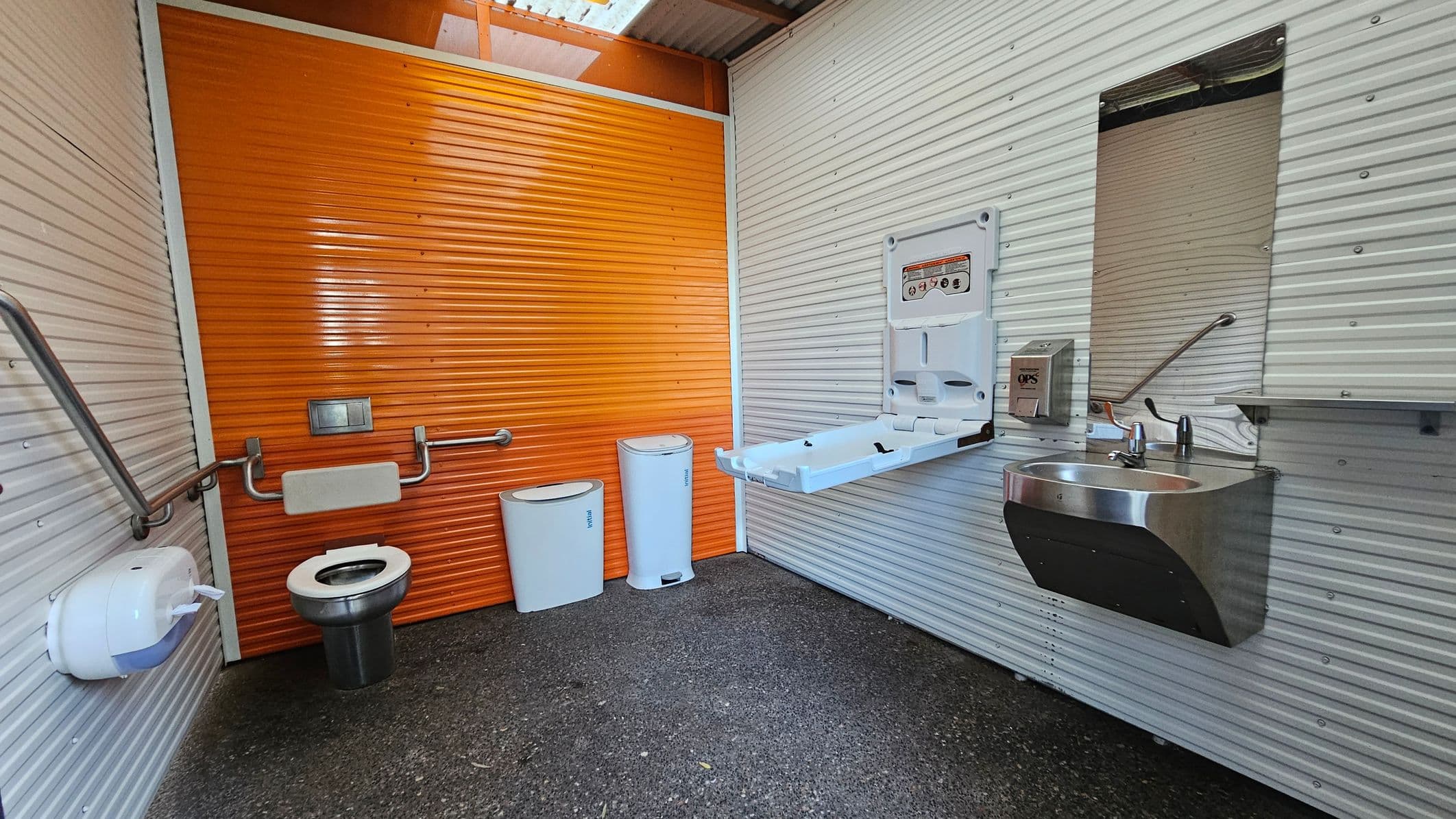 A public restroom with corrugated metal walls, featuring an orange wall at the back. On the left, a stainless steel toilet with grab bars and a toilet paper dispenser. Two white bins are next to the toilet. On the right, a baby changing station is mounted on the wall above a stainless steel sink with a mirror and a soap dispenser. The floor is textured and dark, and the ceiling has a translucent panel allowing natural light.