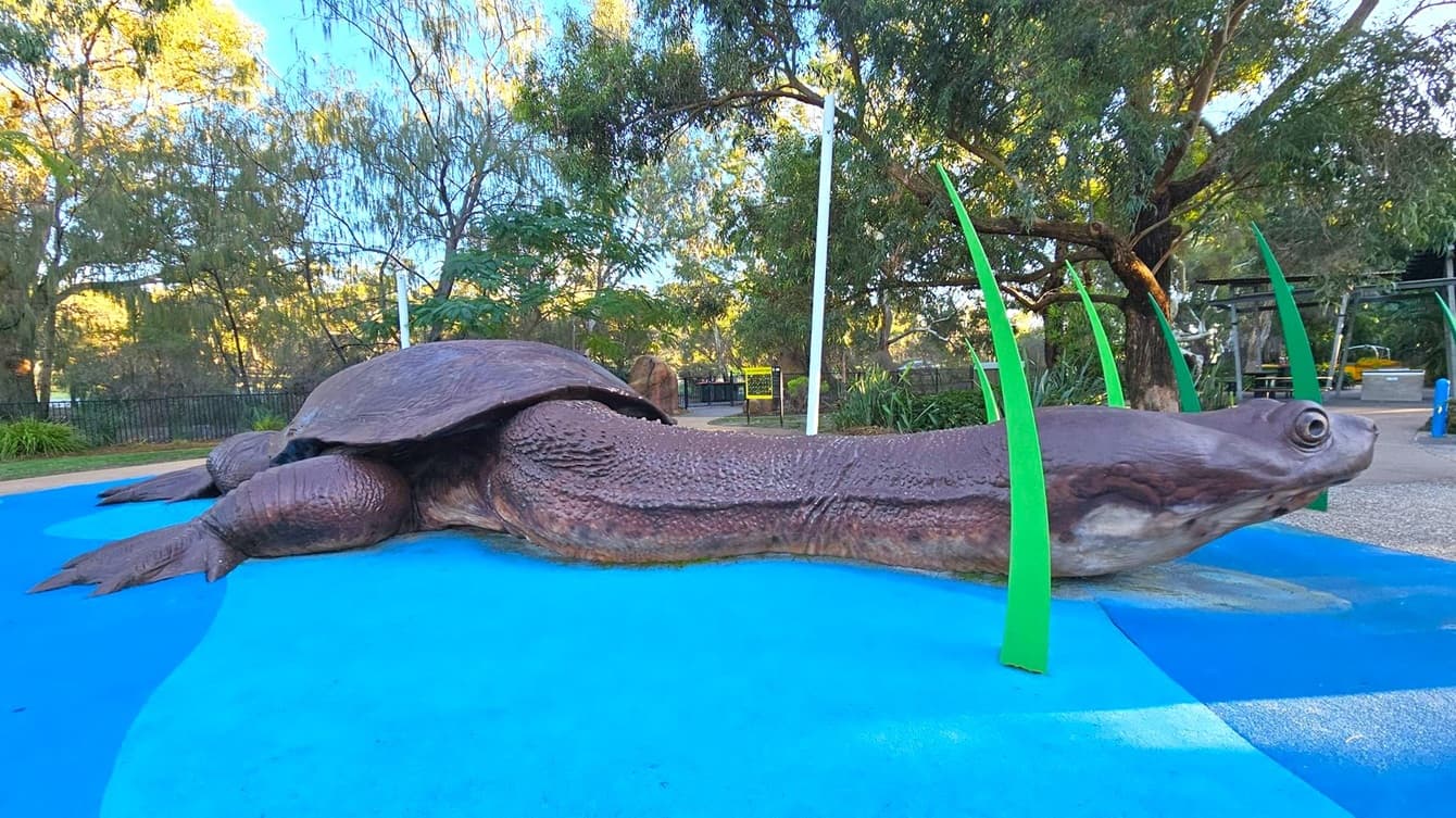 A large sculpture of a turtle is positioned on a blue surface, resembling water. The turtle has a textured shell and extended neck. Green, blade-like structures protrude from the ground around it. In the background, there are trees with dense foliage and a pathway leading to a fenced area. The scene is outdoors, with natural light filtering through the trees.
