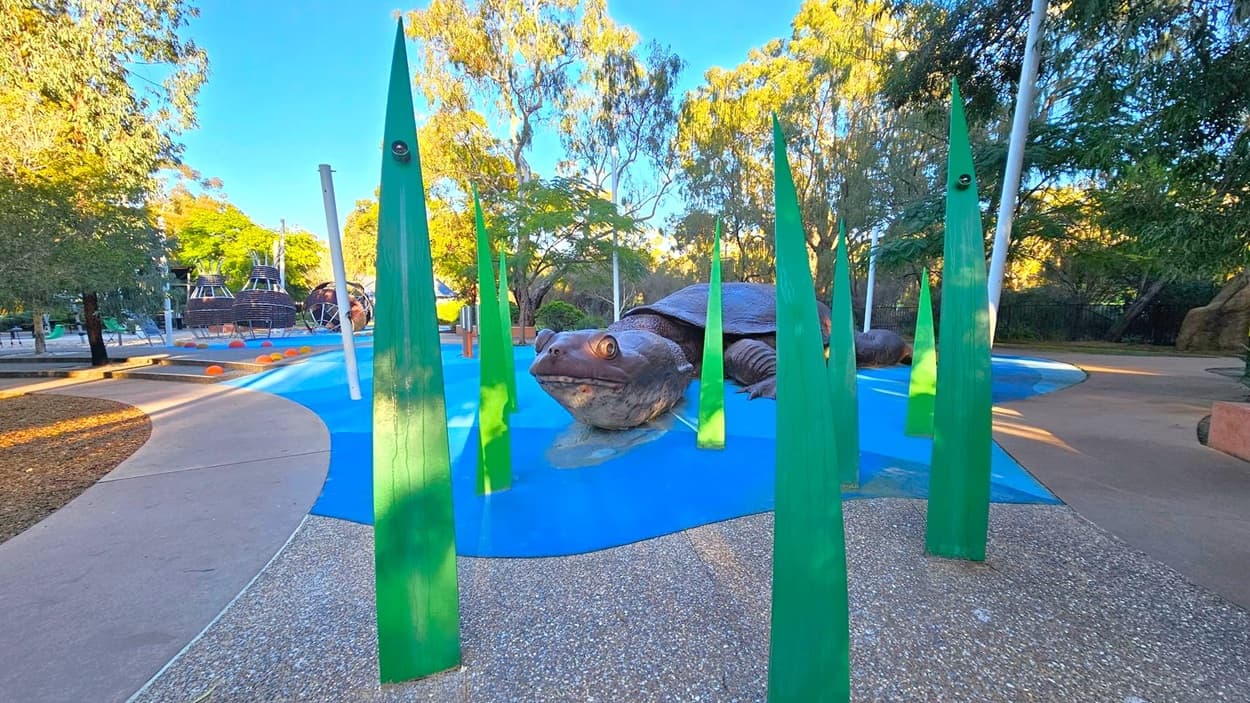 A large turtle sculpture is in the centre, surrounded by tall, green, leaf-like structures. The ground is a mix of blue and speckled concrete, resembling water and land. In the background, there are trees and a playground area with structures resembling woven baskets. The area is well-lit with natural light, and the scene is set in a park with a mix of paved and natural surfaces.