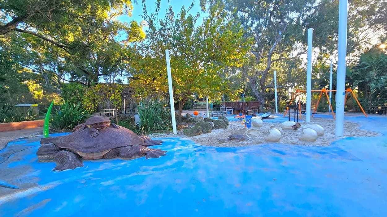 A playground with a large turtle sculpture on the left, set on a blue surface. In the centre, there are white poles and a tree with green leaves. The ground transitions to a sandy area with scattered white spherical objects. On the right, an orange swing set is visible. The background is filled with dense trees and foliage, providing shade and a natural setting.