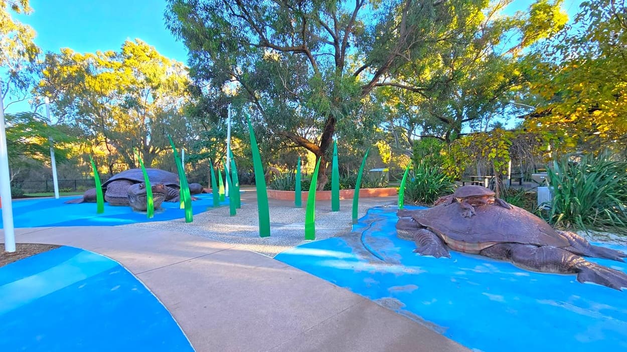 A park scene with two large turtle sculptures on a blue surface resembling water. The left turtle is partially obscured by tall, green, leaf-like structures. The right turtle is more visible, with detailed features. The background is filled with lush trees and greenery, and a pathway curves through the scene. The ground is a mix of blue and beige, and the area is well-lit by natural light.