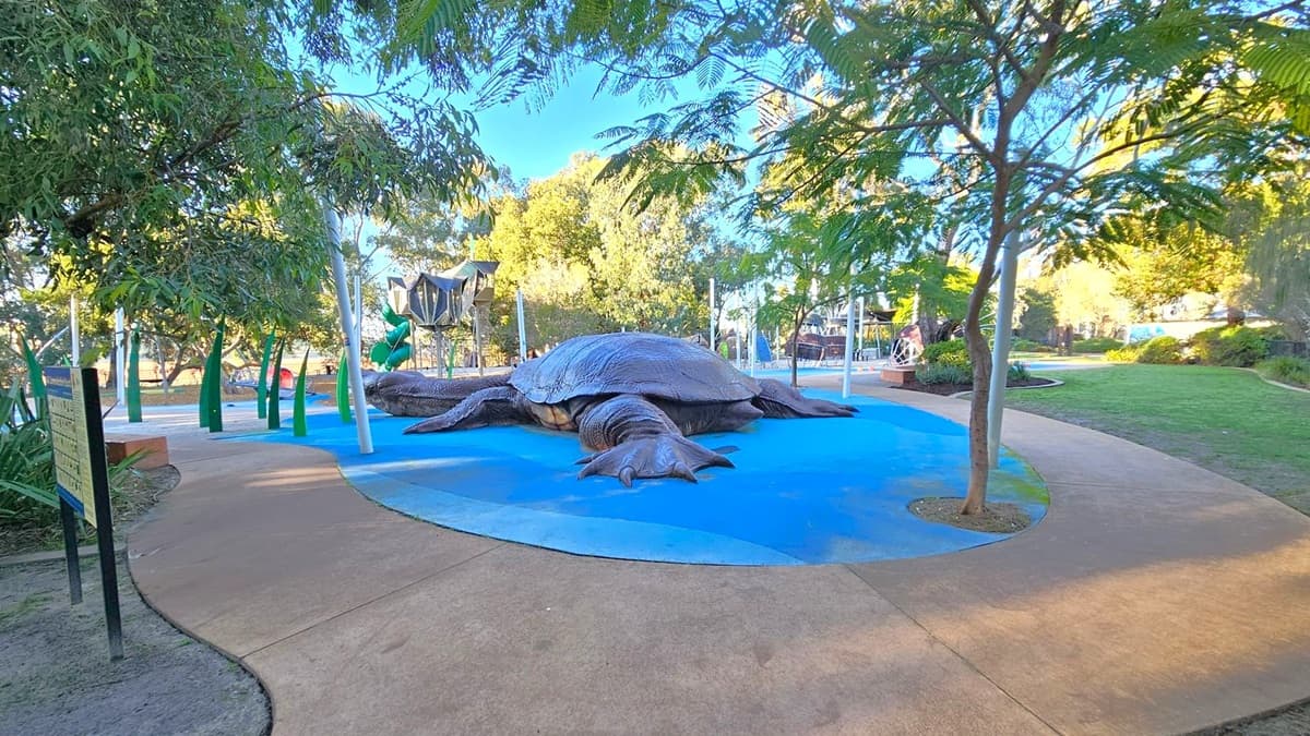 A large turtle sculpture is positioned on a blue surface in the centre of a park. Surrounding the turtle are various playground structures, including slides and climbing frames. Trees with green foliage provide shade, and a curved pathway made of concrete leads through the area. Grass and additional trees are visible in the background, creating a lush, natural setting. A sign is located on the left side near the path.