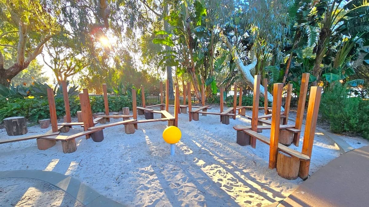 A playground area with wooden structures and balance beams set in sand. In the centre, there's a bright yellow sphere on a post. Tall trees and lush greenery surround the area, with sunlight filtering through the leaves. The ground is sandy, and there is a paved path on the right side.