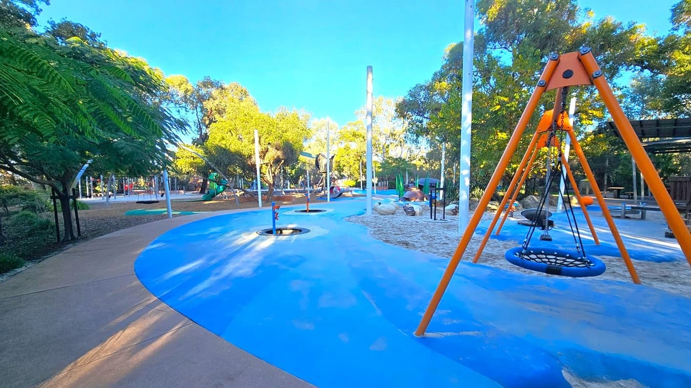 A playground with a blue and brown pathway winding through it. On the right, there's an orange swing set with a circular netted swing. In the background, various play structures and equipment are scattered among trees and greenery. The ground is a mix of sand and rubberised surfaces, and there are several tall white poles. Trees with dense foliage provide shade throughout the area.