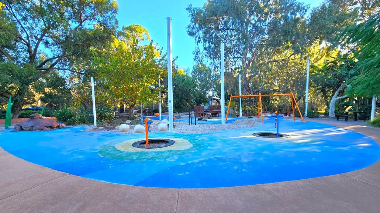 A playground with a blue and green rubber surface. In the centre, there are two circular trampolines and an orange water play feature. To the right, an orange swing set is visible. The background is filled with trees and greenery, providing shade. A sandy area with large white stones is near the centre. The ground is bordered by a concrete path.
