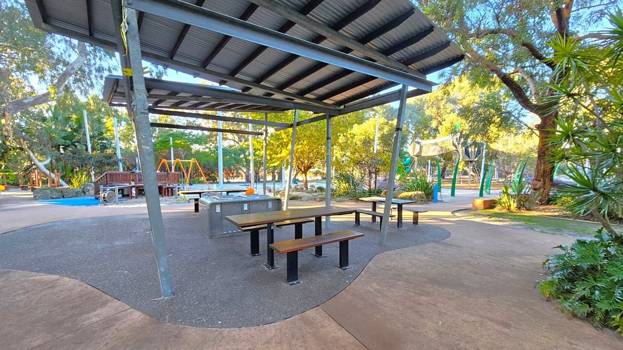 A park setting with a covered picnic area in the foreground, featuring wooden benches and tables on a speckled surface. To the left, there are swings and a wooden play structure. The background is filled with trees and greenery, with a path winding through the park. The ground is a mix of concrete and textured surfaces, and the ceiling of the shelter is made of corrugated metal.