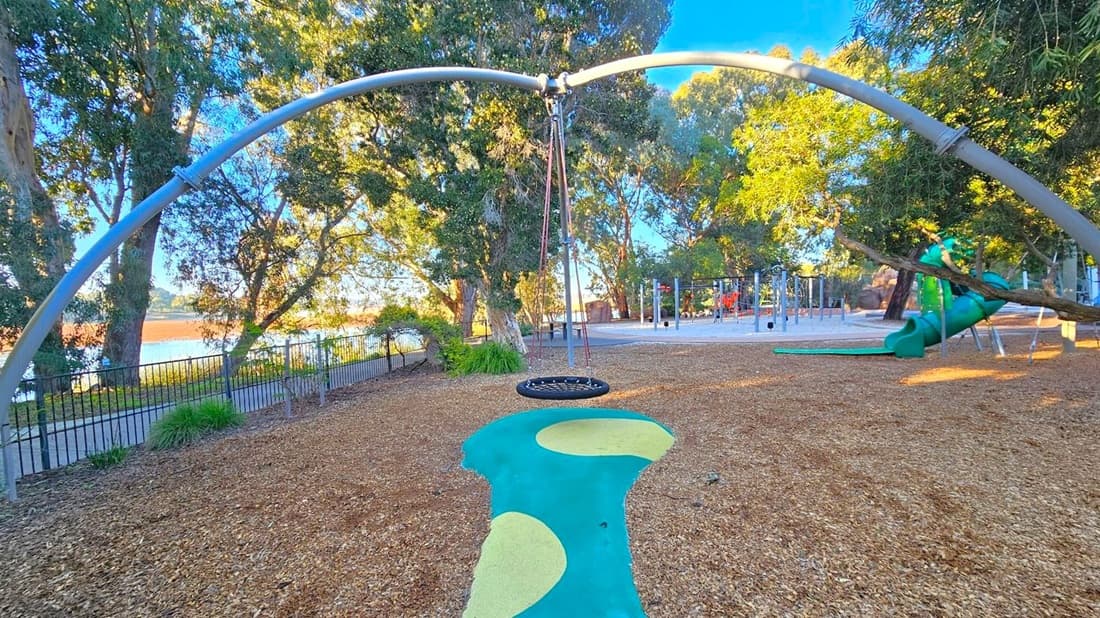 A playground with a large circular swing suspended from a metal arch in the centre. The ground is covered with wood chips, and there's a blue and yellow painted path beneath the swing. To the right, a green slide is visible among trees. In the background, more playground equipment is seen, along with a fence and a body of water. Tall trees surround the area, providing shade.